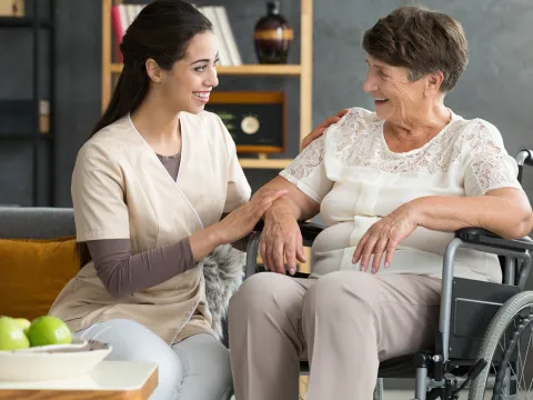 A woman in a wheelchair, with her caretaker sitting next to her and comforting her. Both are smiling.