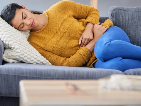 Woman, lying down on a sofa, her knees to her chest, clutching her stomach because she is experiencing endometriosis pain.