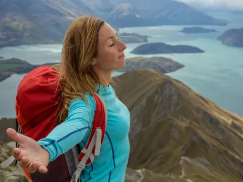 Woman with blue shirt and red pack standing on mountain top, arms spread with mountain range and lake around her