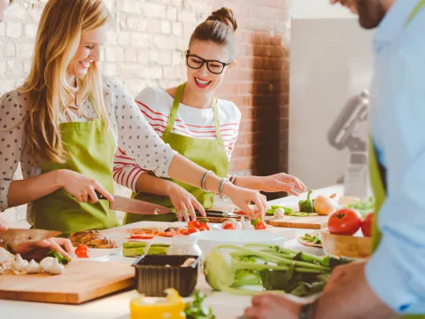 A group of friends are standing around a counter, cutting vegetables at a cooking class.