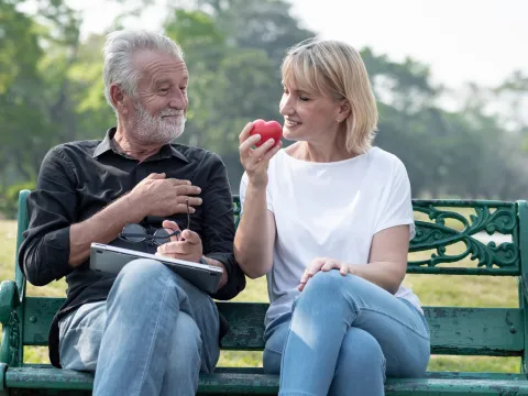 A senior couple sitting on a bench in the park, the woman holding a red heart.