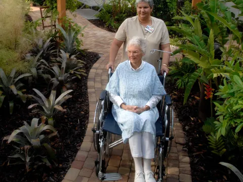 Elder woman in a wheelchair being pushed down a path through a garden.