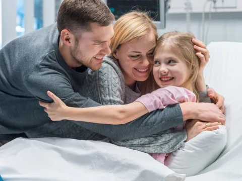 A family hugging their young daughter in her hospital bed.