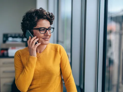 A woman with short, curly hair and glasses in a yellow sweater, on her phone, looking out a window.