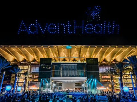 AdventHealth Orchestra performing at a night outdoor concert at Dr. Phillips Center for the Performing Arts with drones making the AdventHealth logo in the sky.