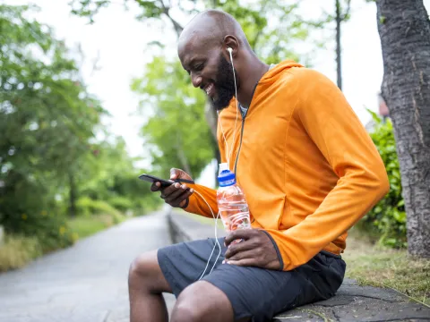 An adult man in an orange hoodie, sitting on a bench outside, looking at his phone and hydrating with a bottle of water.