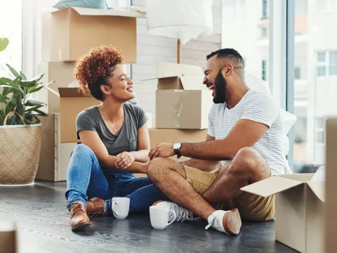 Couple sits on the floor, laughing together in their new apartment, surrounded by mountains of moving boxes.