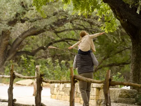 A dad walks through the trees with his daughter on his shoulders