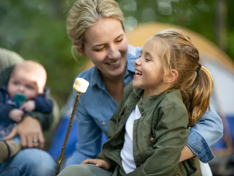 A family of four roasts marshmallows at a campground, the mother is holding her daughter as they smile.