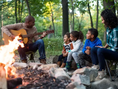 A family singing along while the father plays the guitar around the fireplace in the forest.