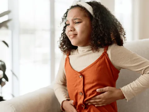 A young girl holding the left side of her abdomen in discomfort while sitting at home.