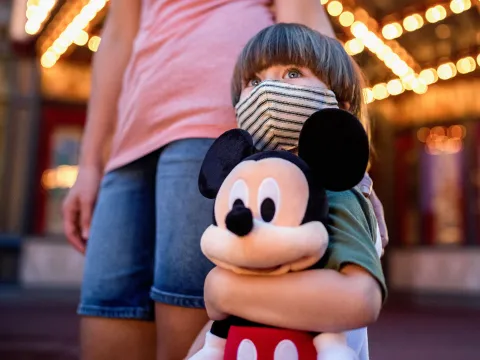 Little boy at Disney in a mask holding a stuffed Mickey Mouse.
