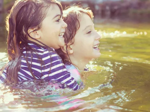 Two young children, one holding the other, swimming in a lake.