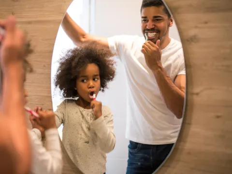 A father and daughter brush their teeth looking in a mirror.