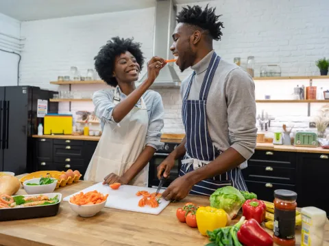 A couple snacking on veggies while cooking in the kitchen.