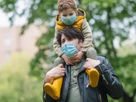 A father carrying his son on his shoulders while wearing a mask. 