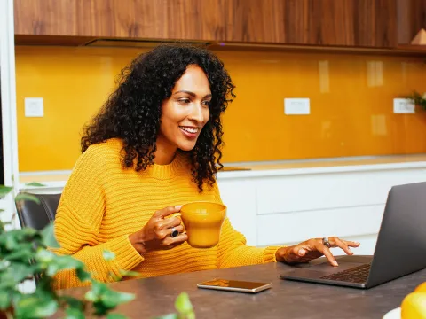 A woman in her kitchen and drinking coffee while on her laptop