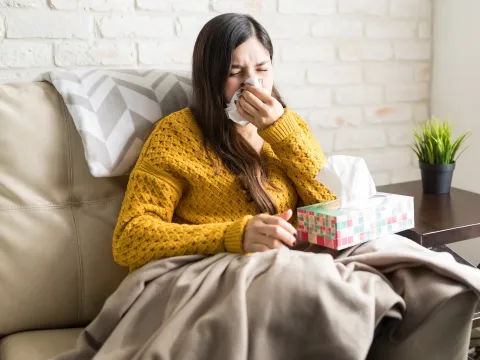 A young woman sitting on her couch sneezing. 