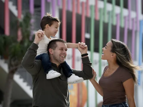 A child riding on his fathers shoulders while his smiling mother walks along side.