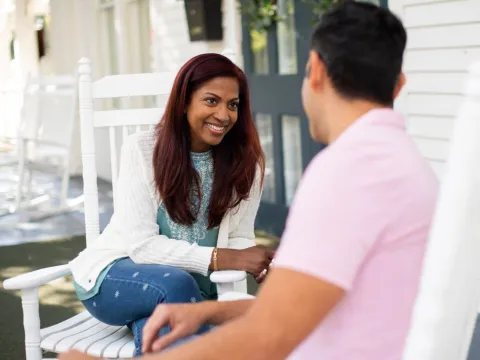A young hispanic couple sit in rocking chairs on the porch together.