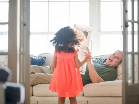 Father laying on sofa reading newspaper with his daughter. 