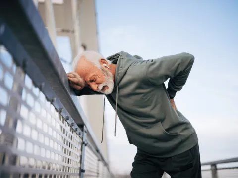 A senior man hunching over against a fence with his hand resting on his back in pain.