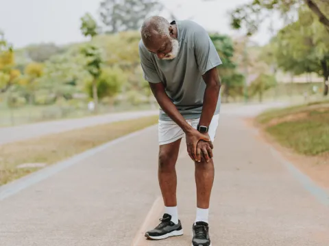 A senior black man takes a break from exercising outside and places his hands on his left knee.