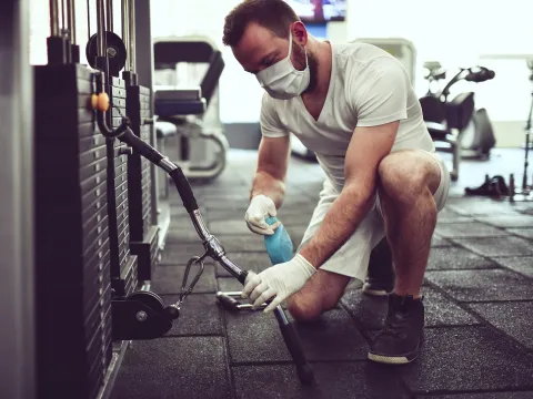 A man wearing a mask and cleaning fitness equipment in a gym.