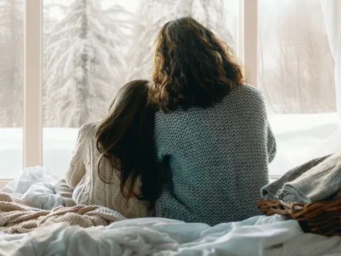 Mother and daughter looking through a window at the snow outside.