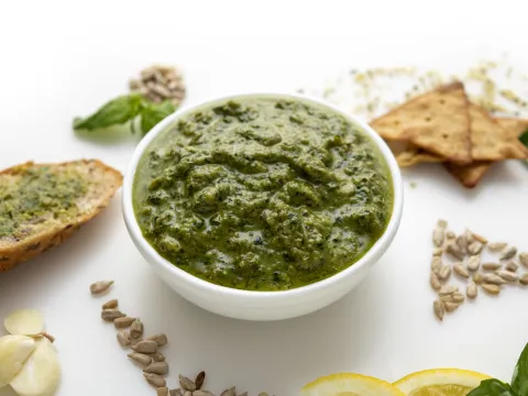 White bowl of green pesto surrounded by a piece of toast and a few crackers.