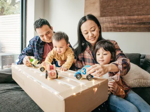 Parents and two children playing with wooden cars on top of a cardboard box while sitting on the couch at home.