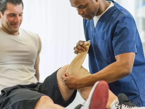 A physical therapist bandaging a male patient's left knee.