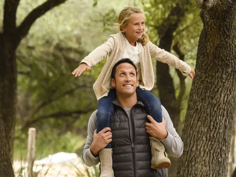 A Caucasian father carries his daughter on his shoulders while on a hike.