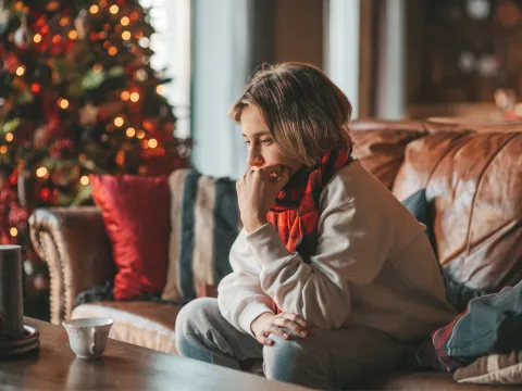 A teen boy sits on a couch at home looking introspectively near a Christmas tree.