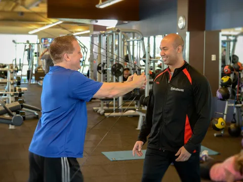 A Caucasian man exercises with a machine under the watch of his trainer.