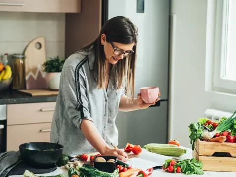 A woman writing down a recipe while in the kitchen and a lot of produce is on the counter.