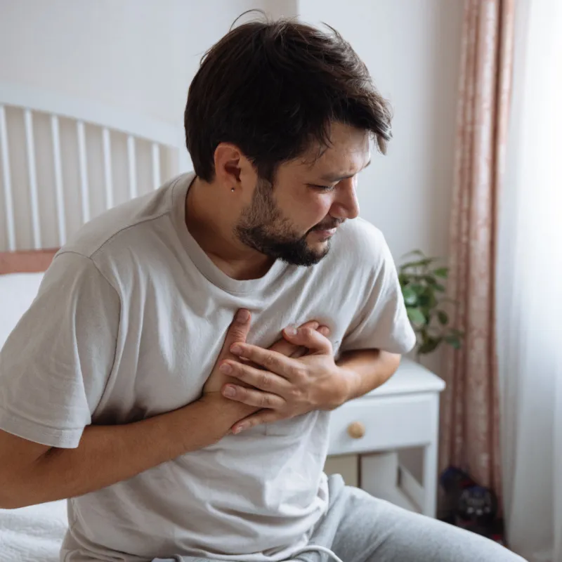 A Man Holds his Hands to his Chest While Sitting on his Bed.