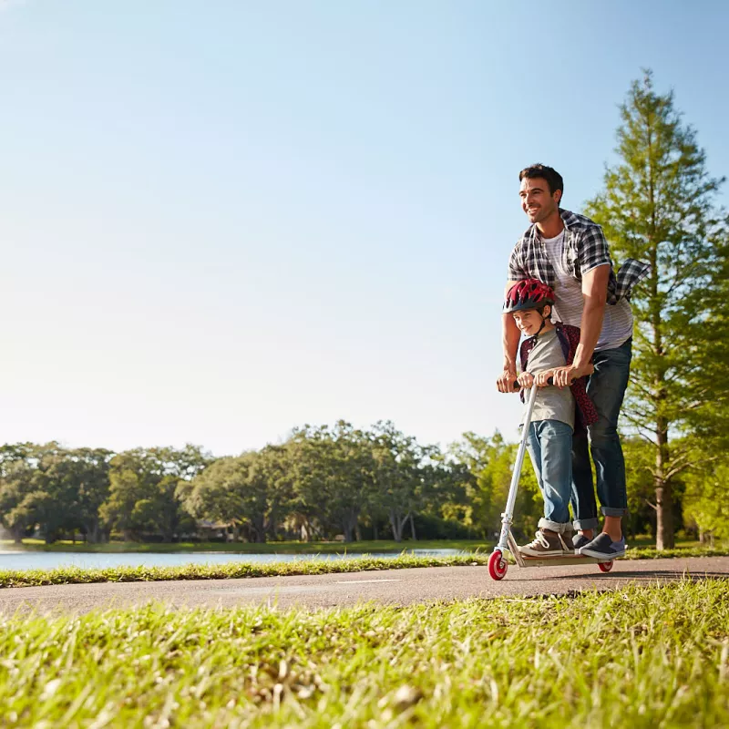 A dad rides a scooter with his son on a sunny day in the park. 