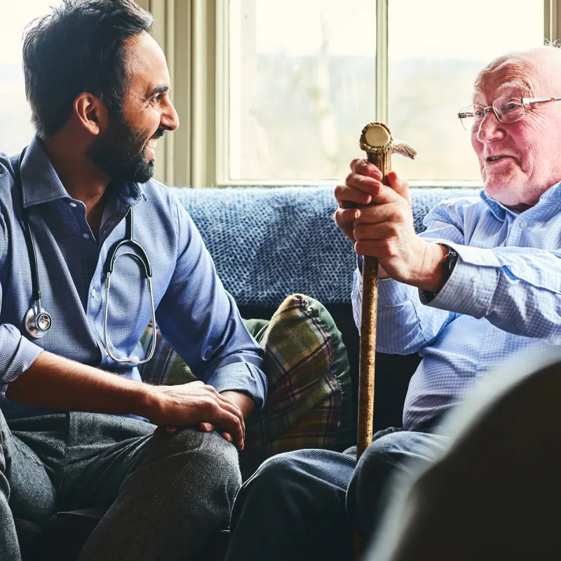 A doctor, sitting on a sofa with a senior man holding a cane, making a home visit.