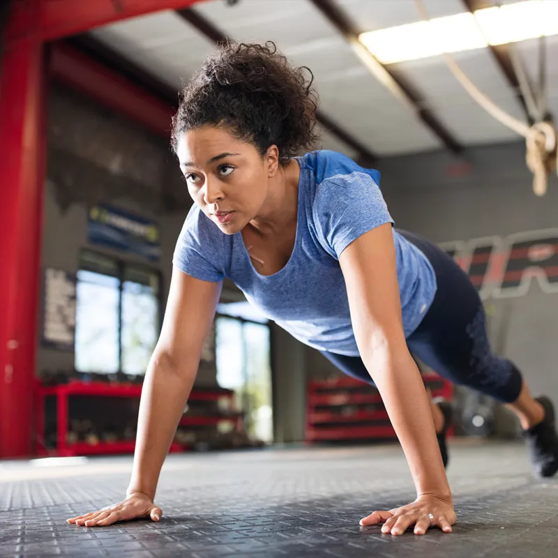 A young African American woman with a blue shirt performs push-ups at an open spaced gym.