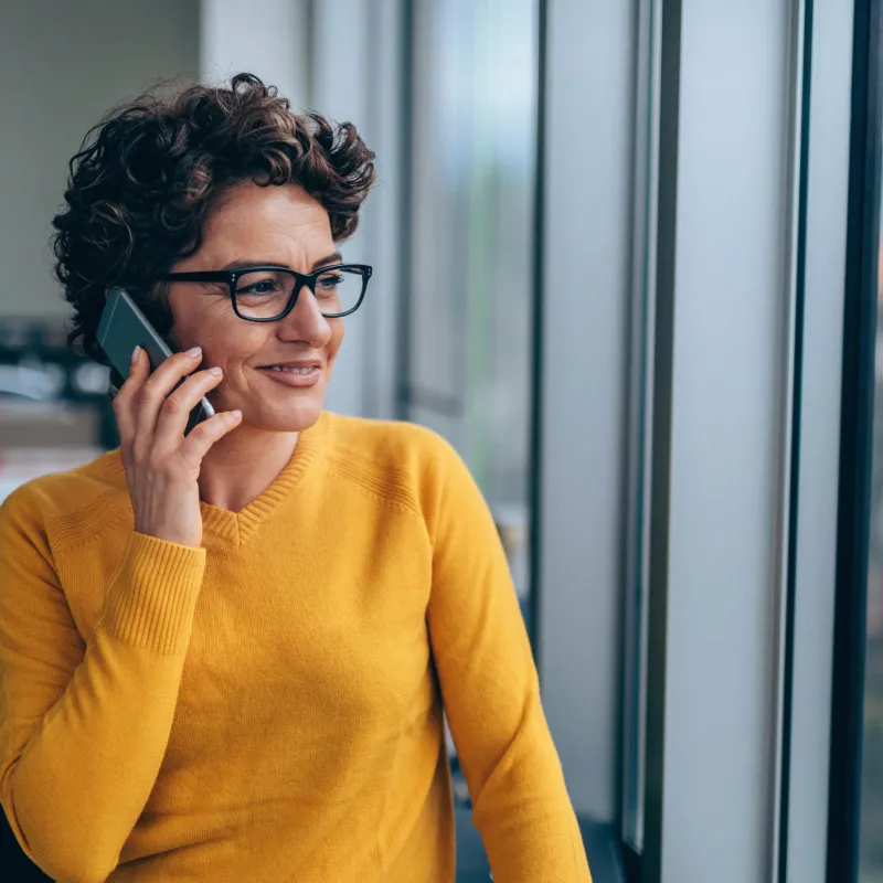 A woman with short, curly hair and glasses in a yellow sweater, on her phone, looking out a window.