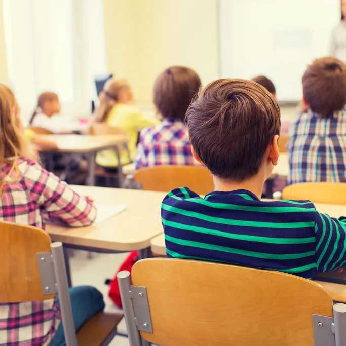 Image of the back of a classroom with several children sitting at their desks facing their teacher and a blackboard.