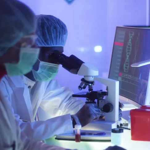 2 individuals in full ppe in a research lab face computers, one looks into a microscope