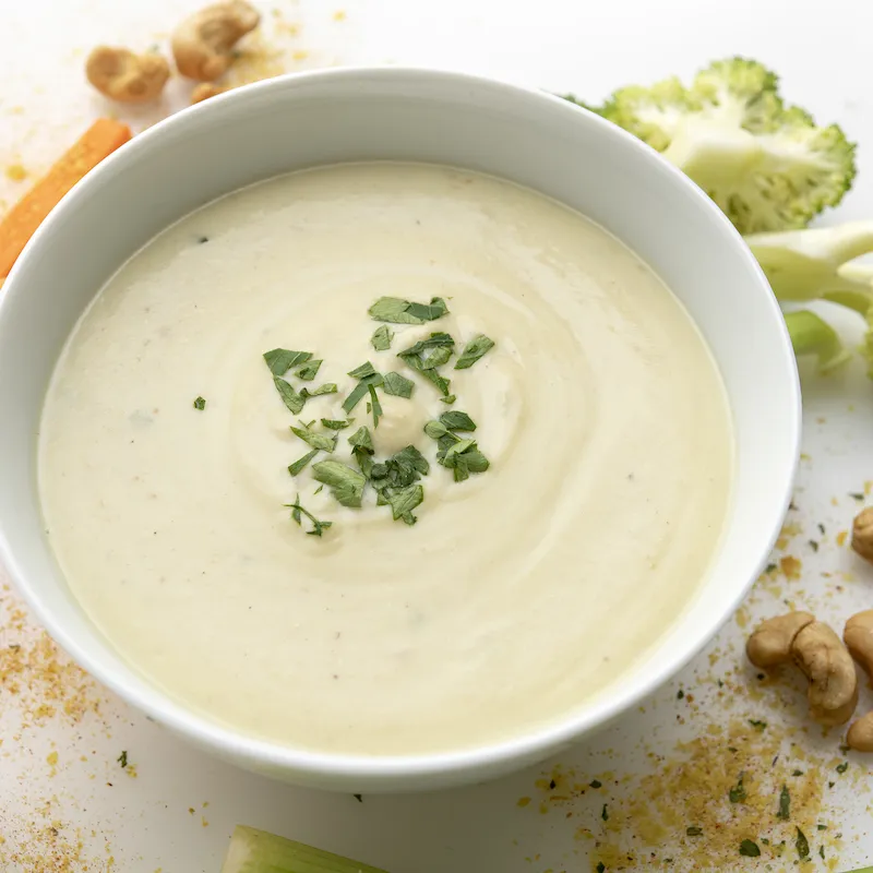 White bowl of cream soup next to carrot sticks, broccoli, and cashews