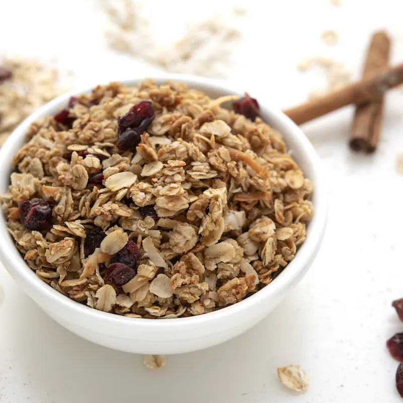 White bowl of granola on white surface with cinnamon stick garnish