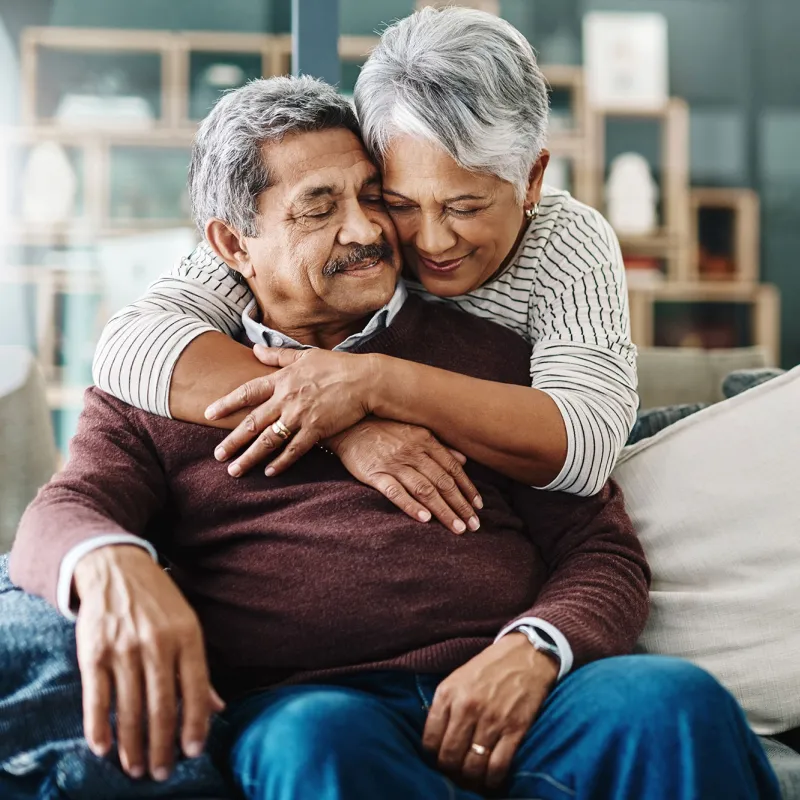 The wife of an old couple hugs her husband on the couch