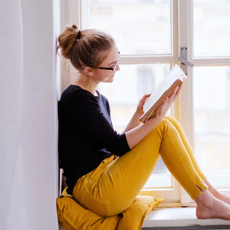 A young woman reads by a window in her home