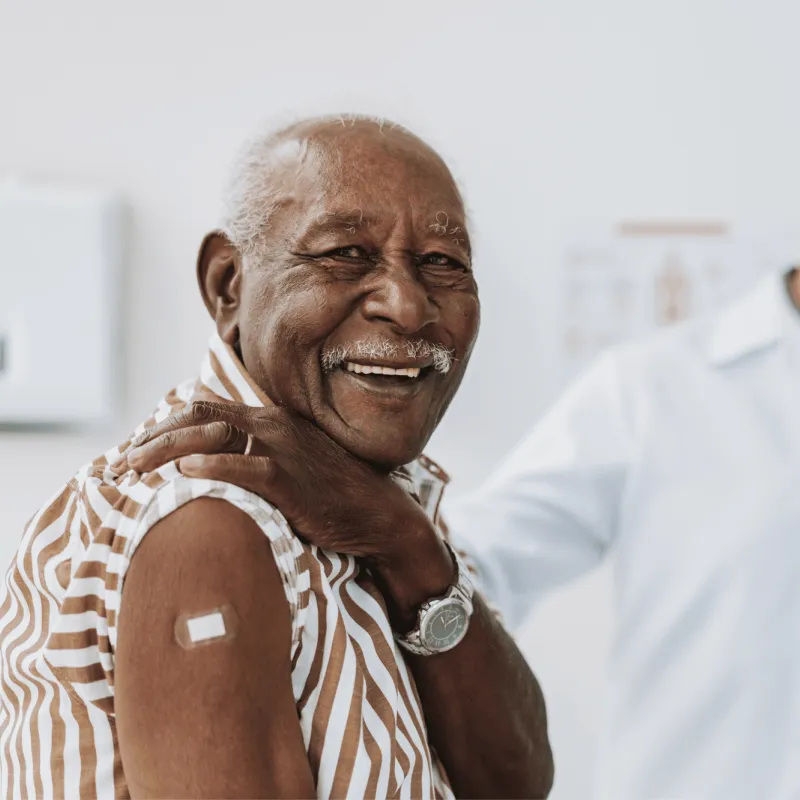 A senior man smiles and shows his arm with bandage after receiving a vaccine in the foreground while a doctor smiles in the background.