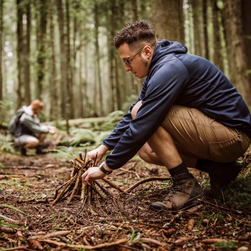 Man building a campfire in a forest while a woman collects timber in the background.