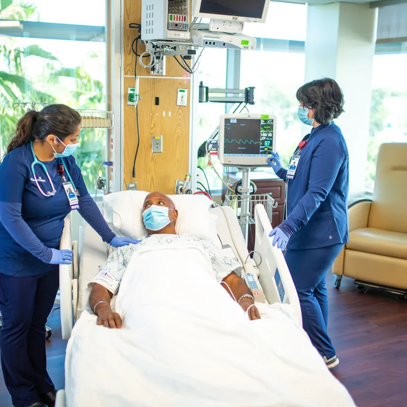 Man in the ICU talking to a nurse while another nurse checks a monitor. All are wearing masks.